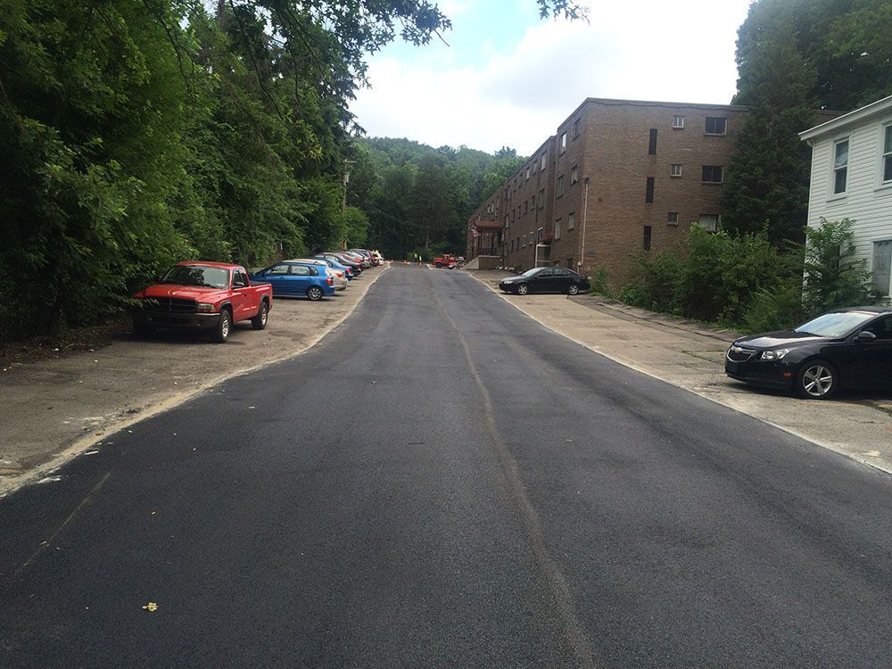 Paved street lined with parked cars and a tall apartment building on a sunny day.
