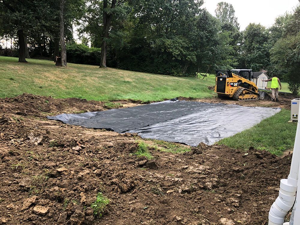Construction of a pond, soil excavation, black fabric lining, a small loader, two workers, and a grassy hill in background.