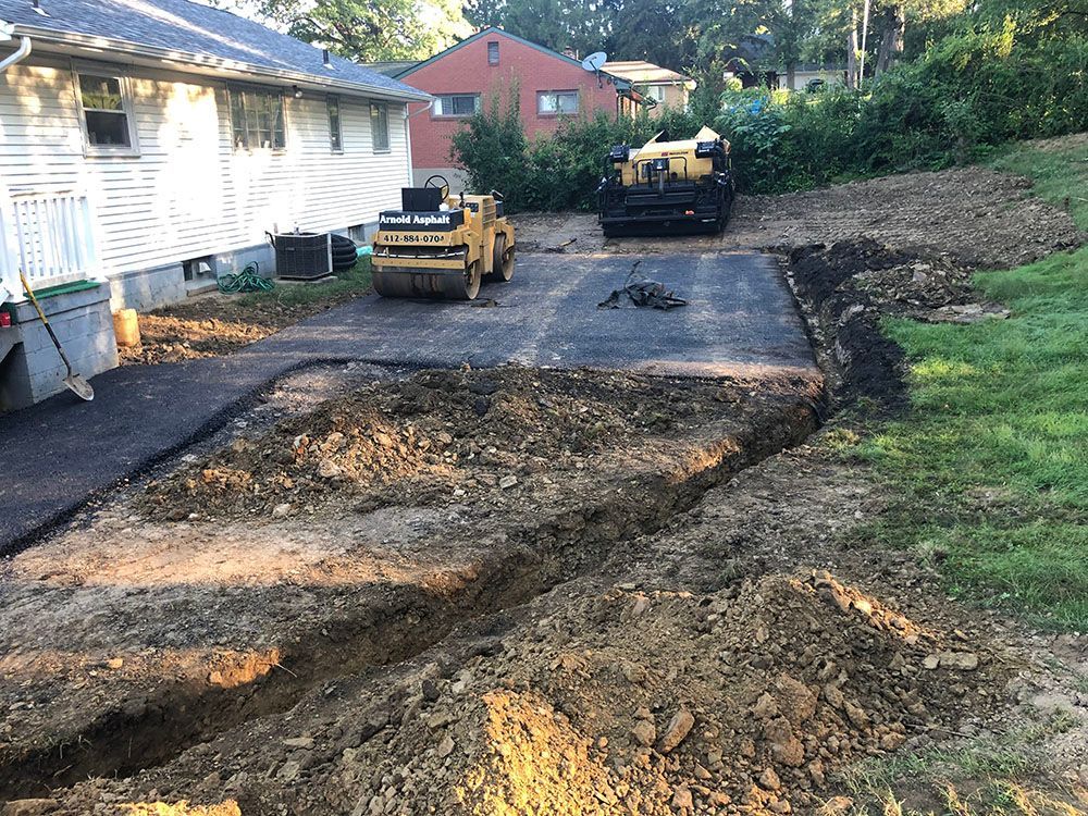 Driveway construction site. Paved area with roller, asphalt machine, and excavated trenches next to a house and grassy area.
