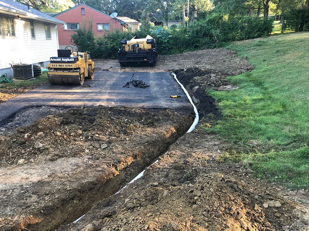 Construction site with asphalt driveway, two rollers, and trench for drainage pipe.