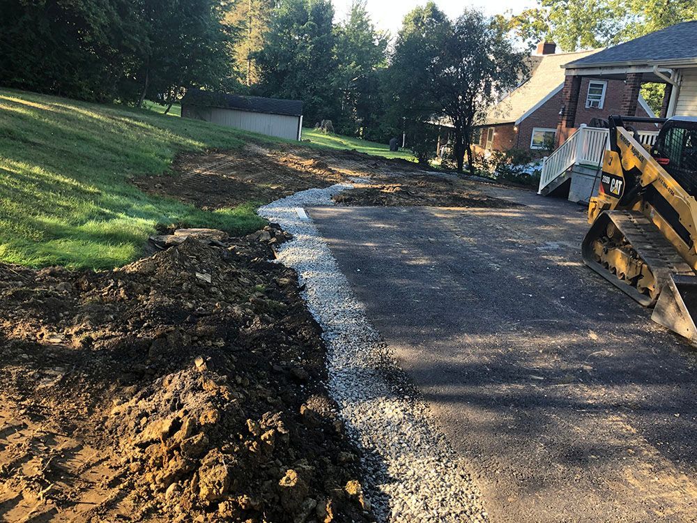 A paved driveway under construction, with a gravel edge, grass slope, and a small excavator.