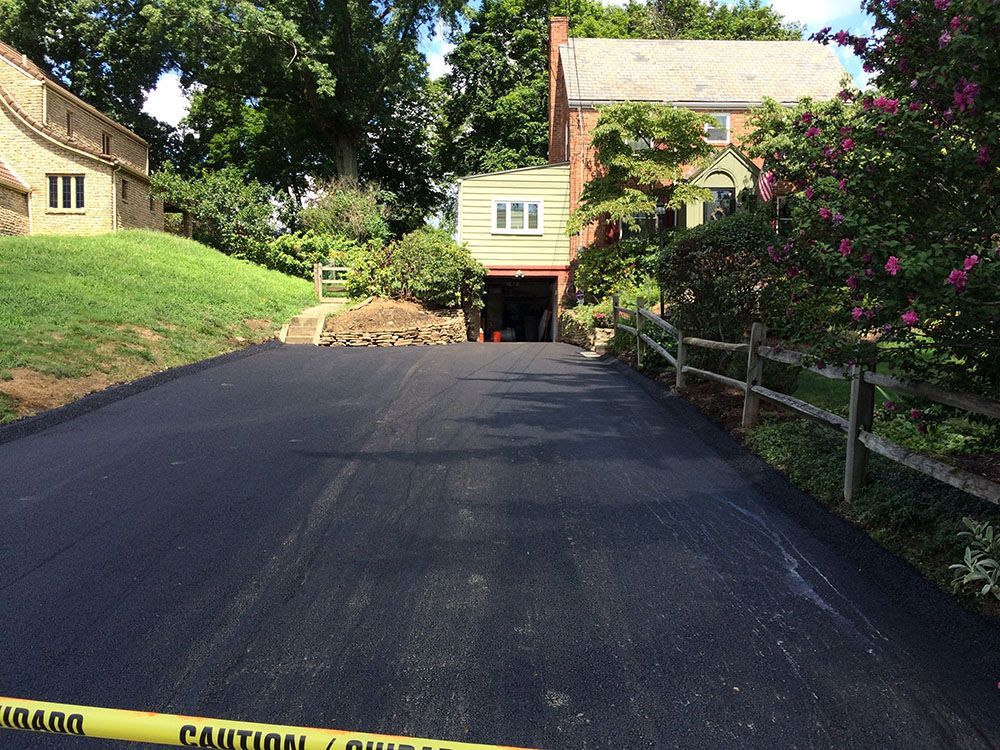 Freshly paved asphalt driveway leading to a garage, with houses and a split-rail fence in a sunny residential setting.