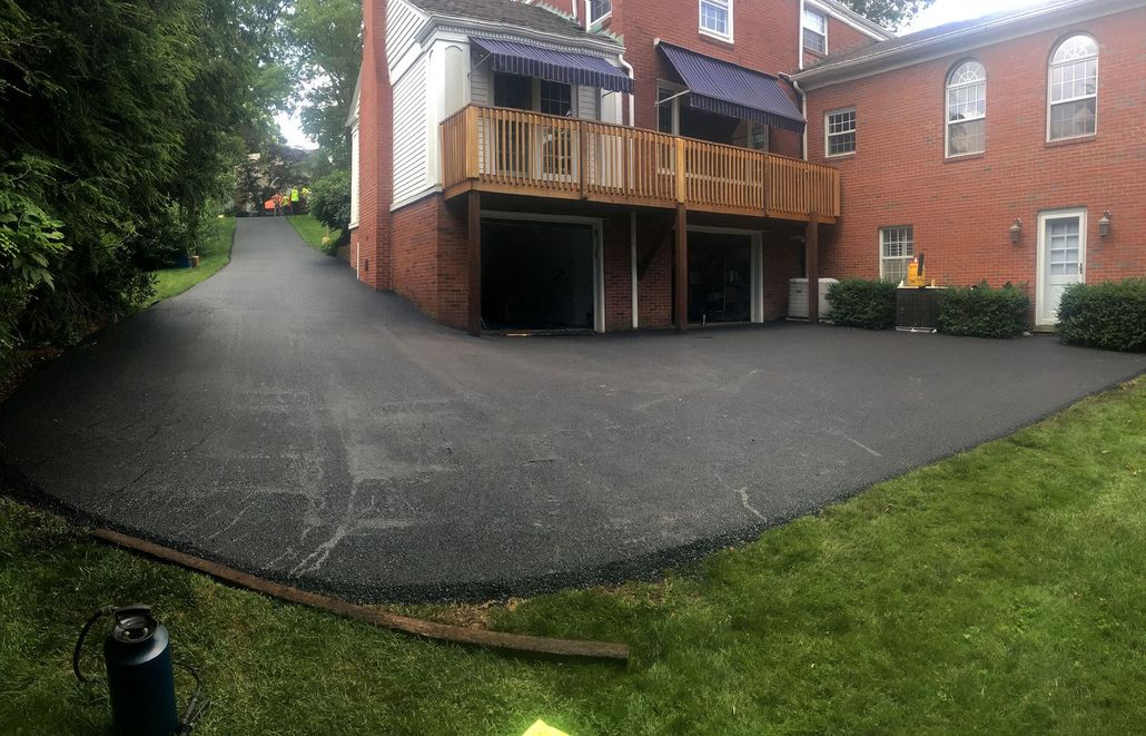 Newly paved black asphalt driveway curves uphill towards houses on a sunny day.