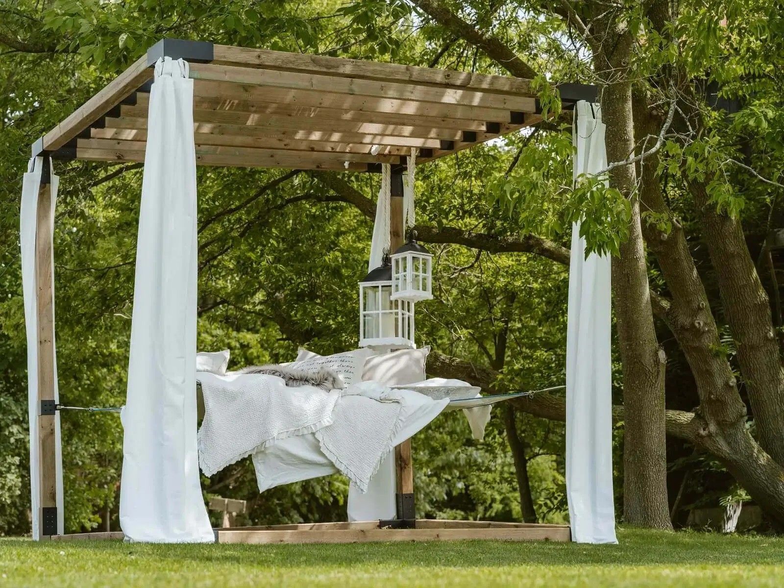 A hammock is sitting under a wooden pergola with white curtains.