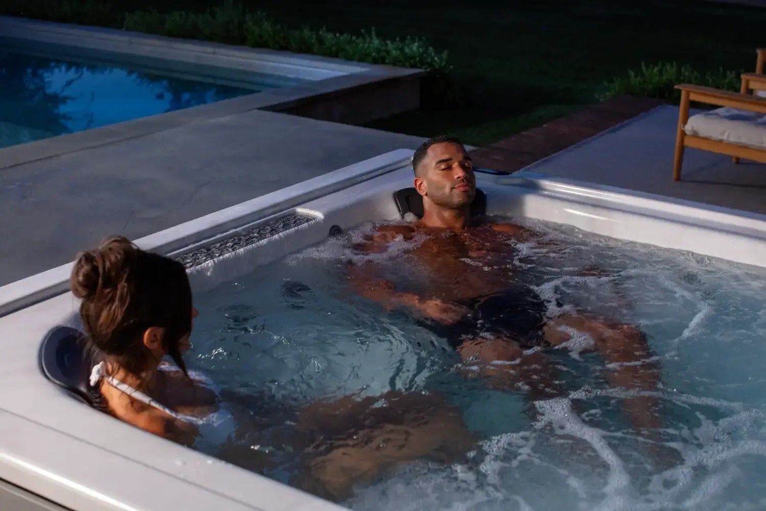 A man and a woman are laying in a hot tub next to a swimming pool.