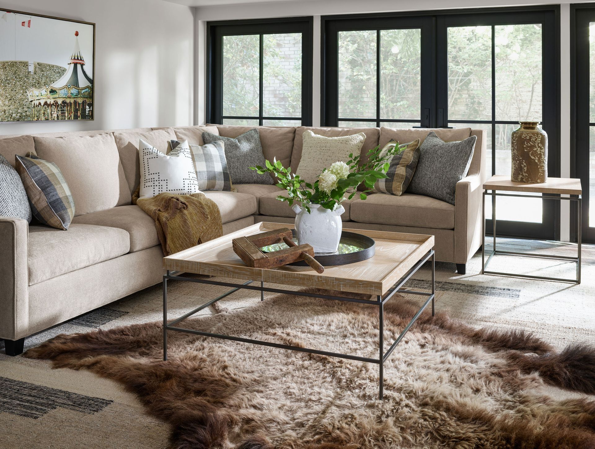 Bright living room with beige sectional, cowhide rug, glass coffee table, and large windows with greenery outside