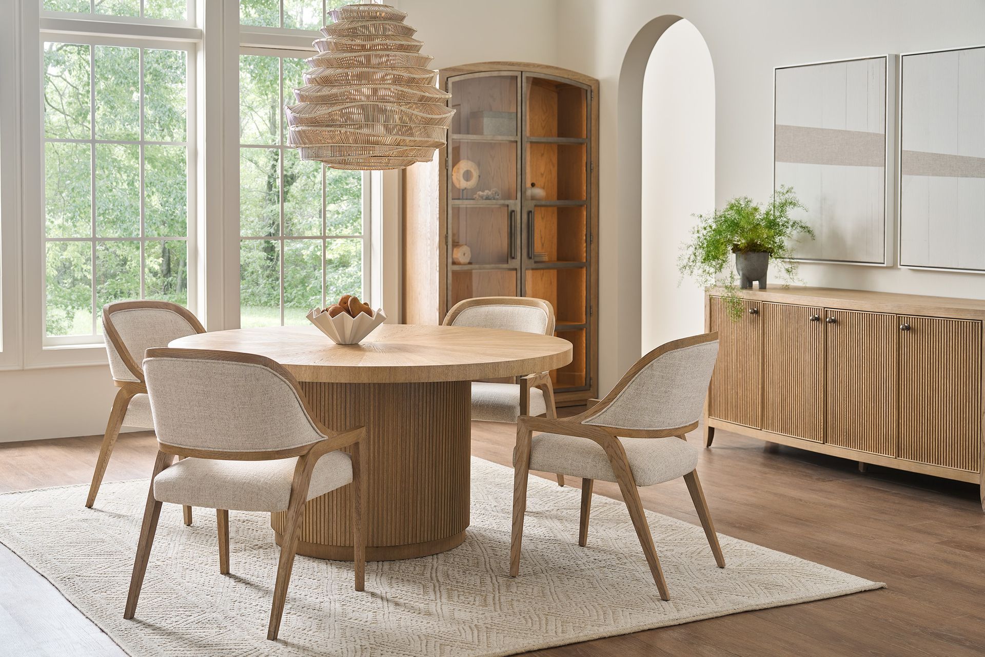 Bright dining room with round wooden table, woven chairs, large windows, and a sideboard with plants