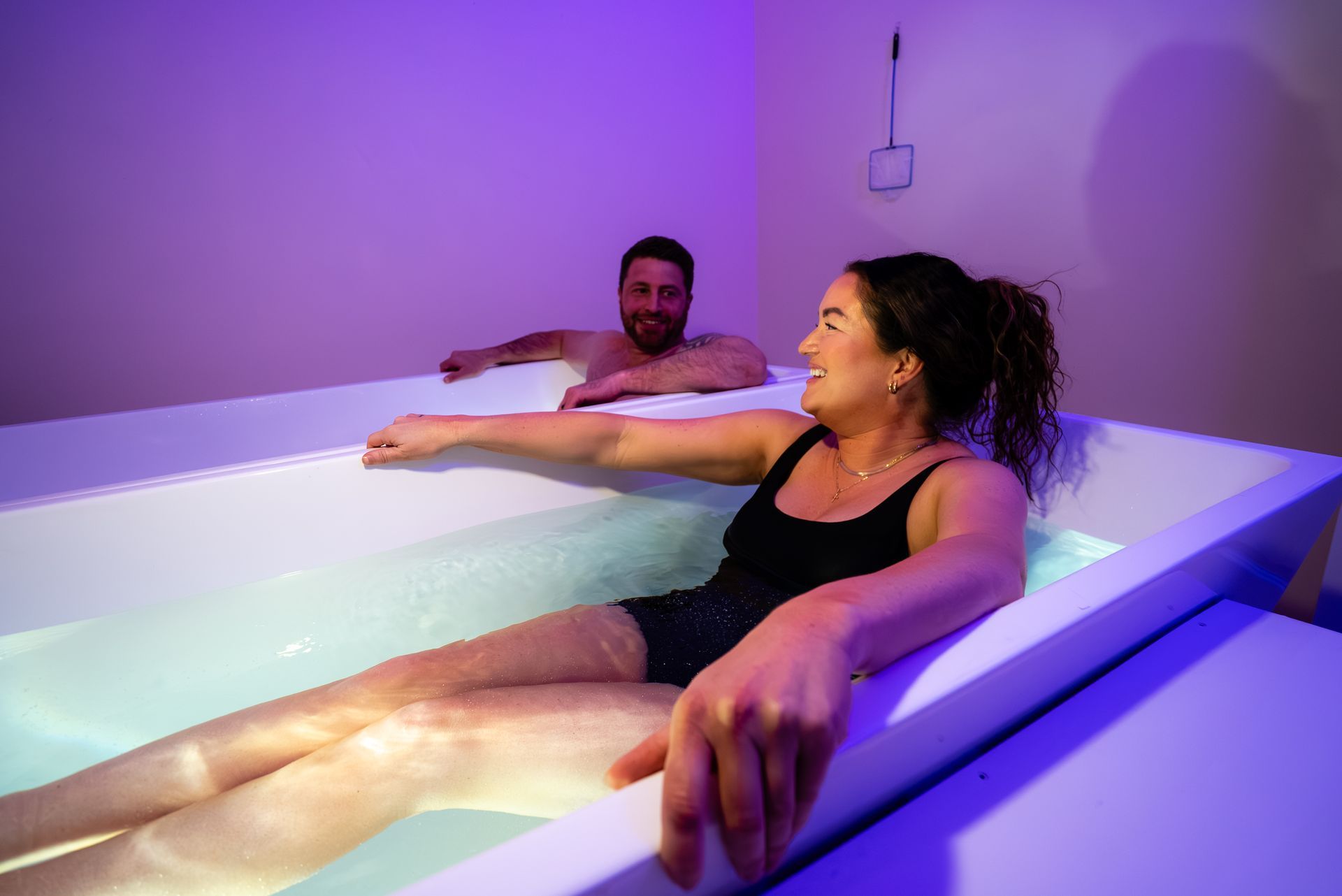 A woman and a man relaxing in a flotation tank with dim purple lighting