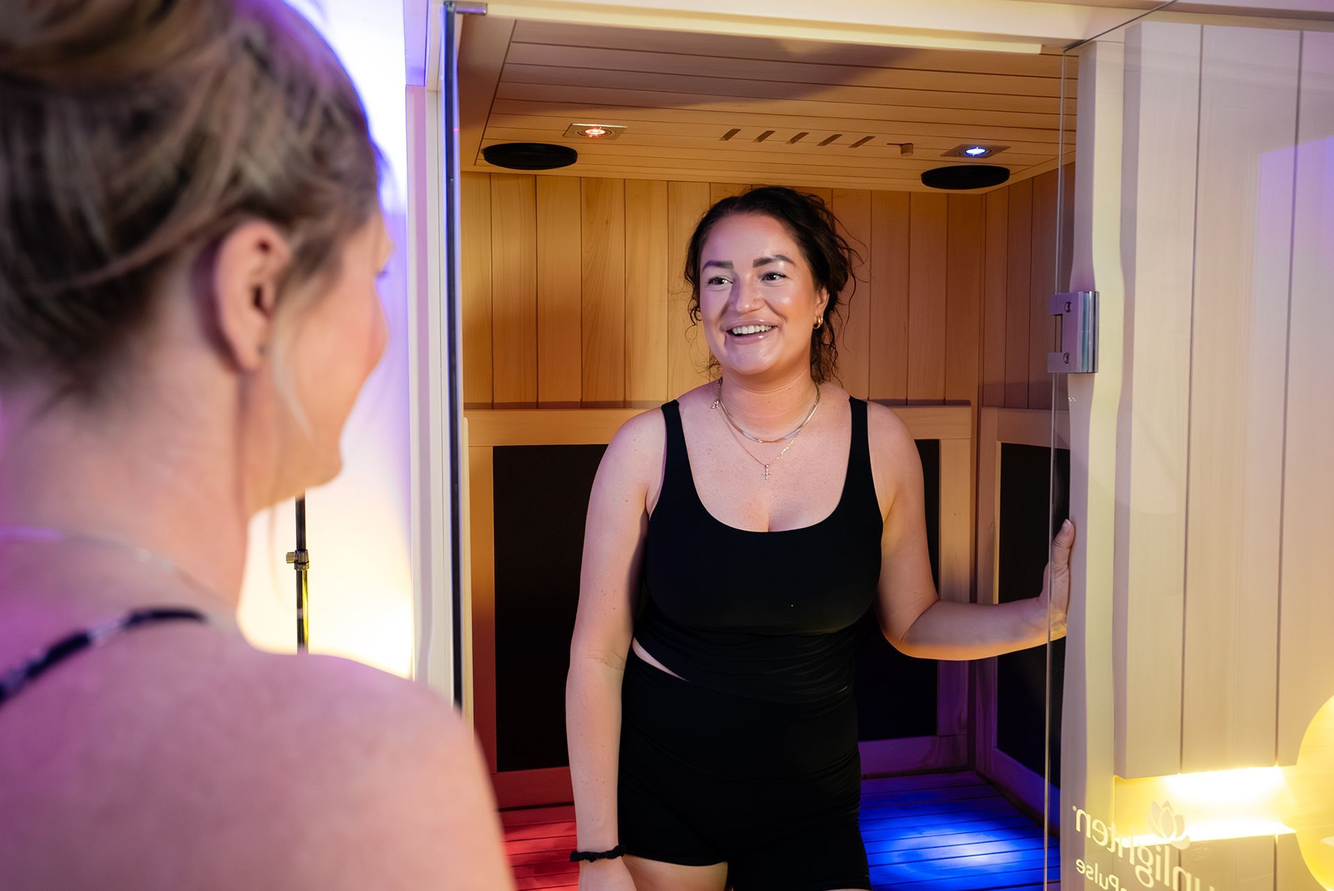 Woman in a black stands in an infrared sauna, smiling and talking to another woman