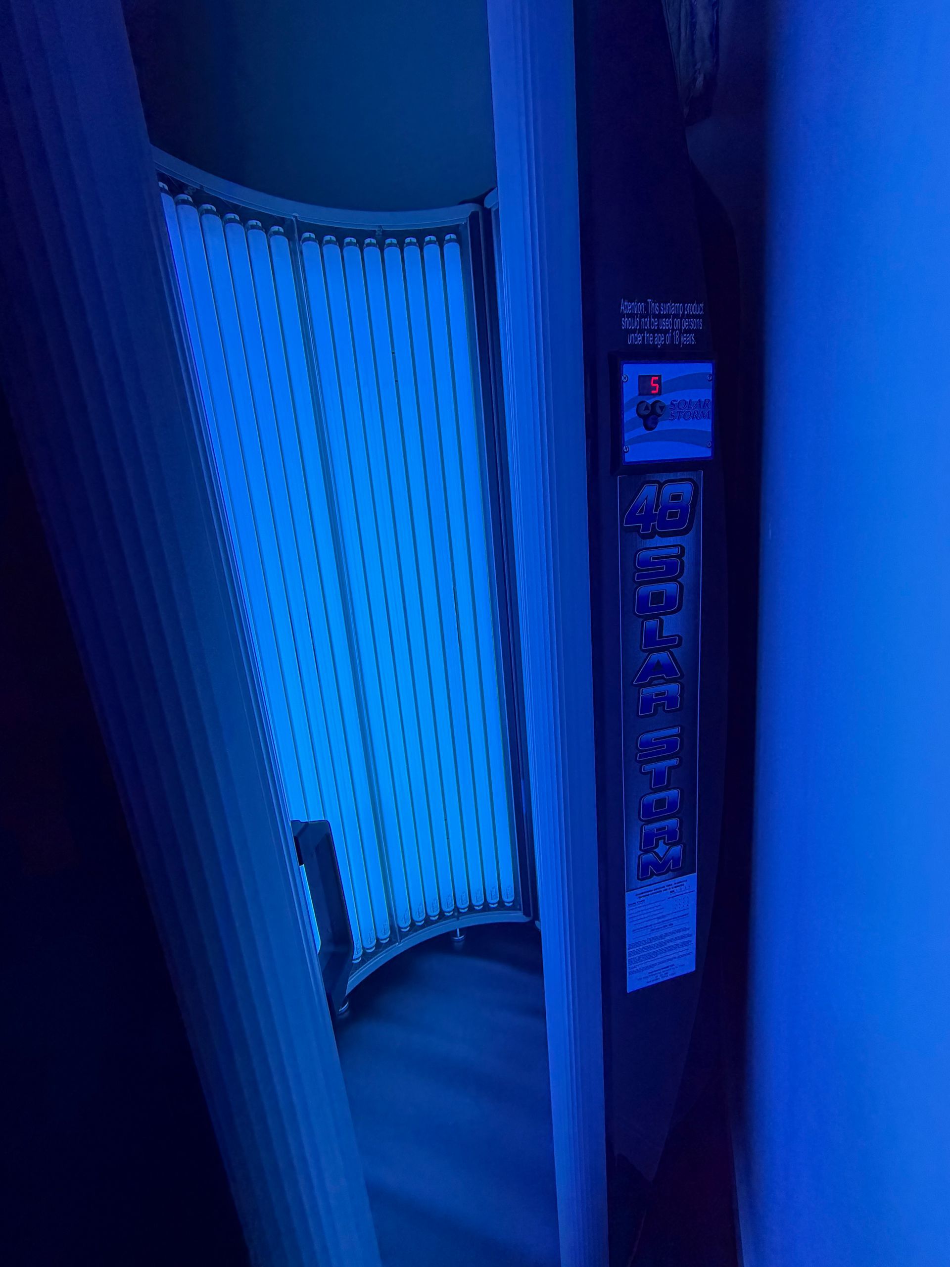 Blue-lit tanning bed booth with an open door and control panel on the side