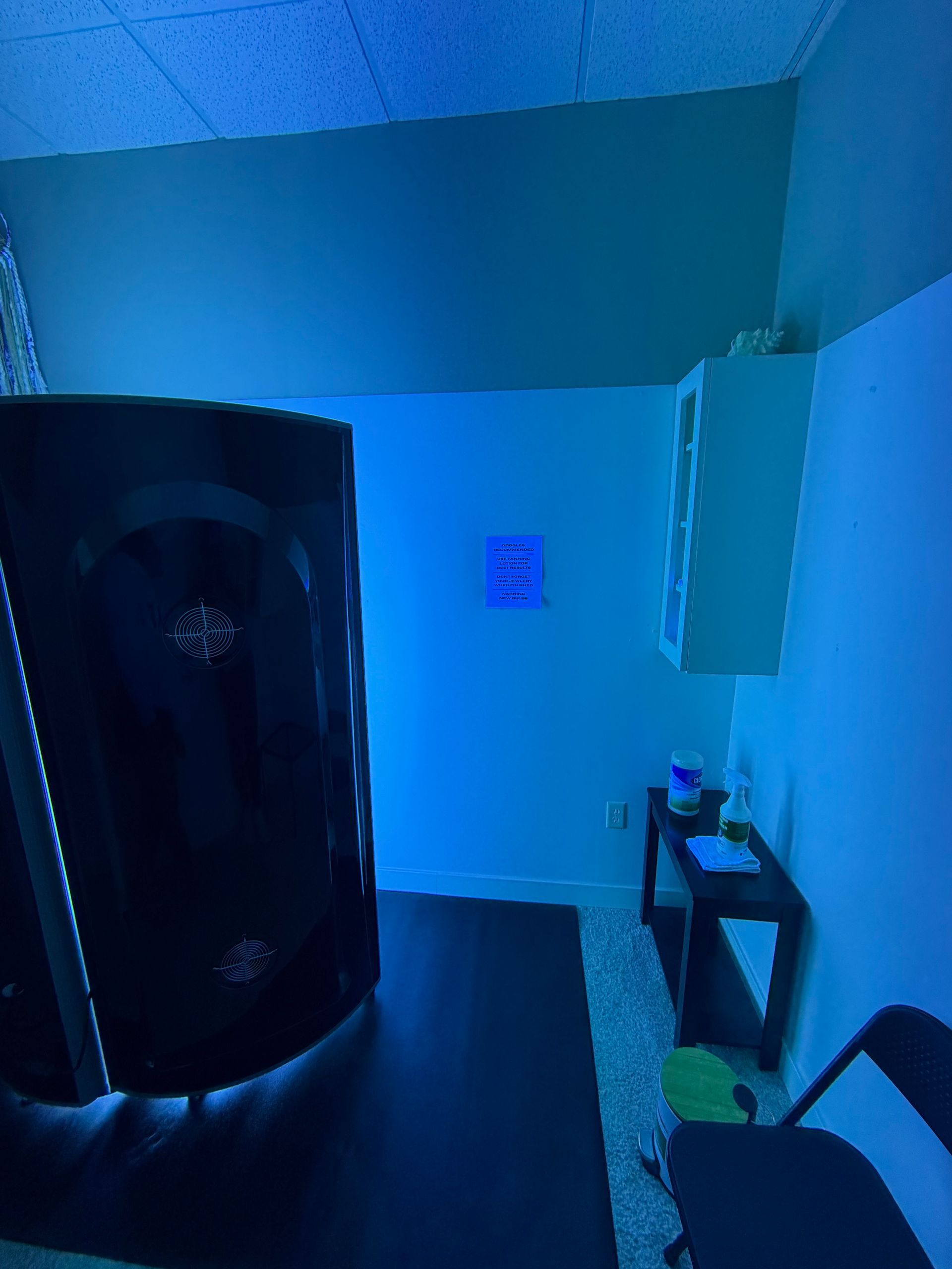 Blue-lit room with a black cylindrical sauna or massage pod, wall cabinet, small shelf, and chair.