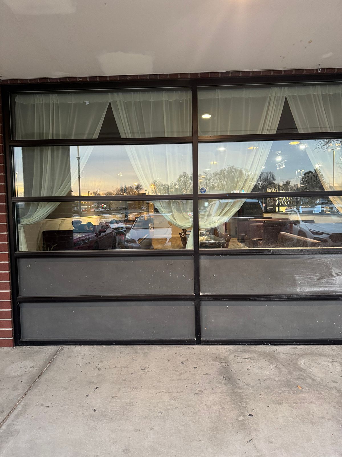 Large glass windows with curtains, reflecting an outdoor scene; brick facade and concrete sidewalk.