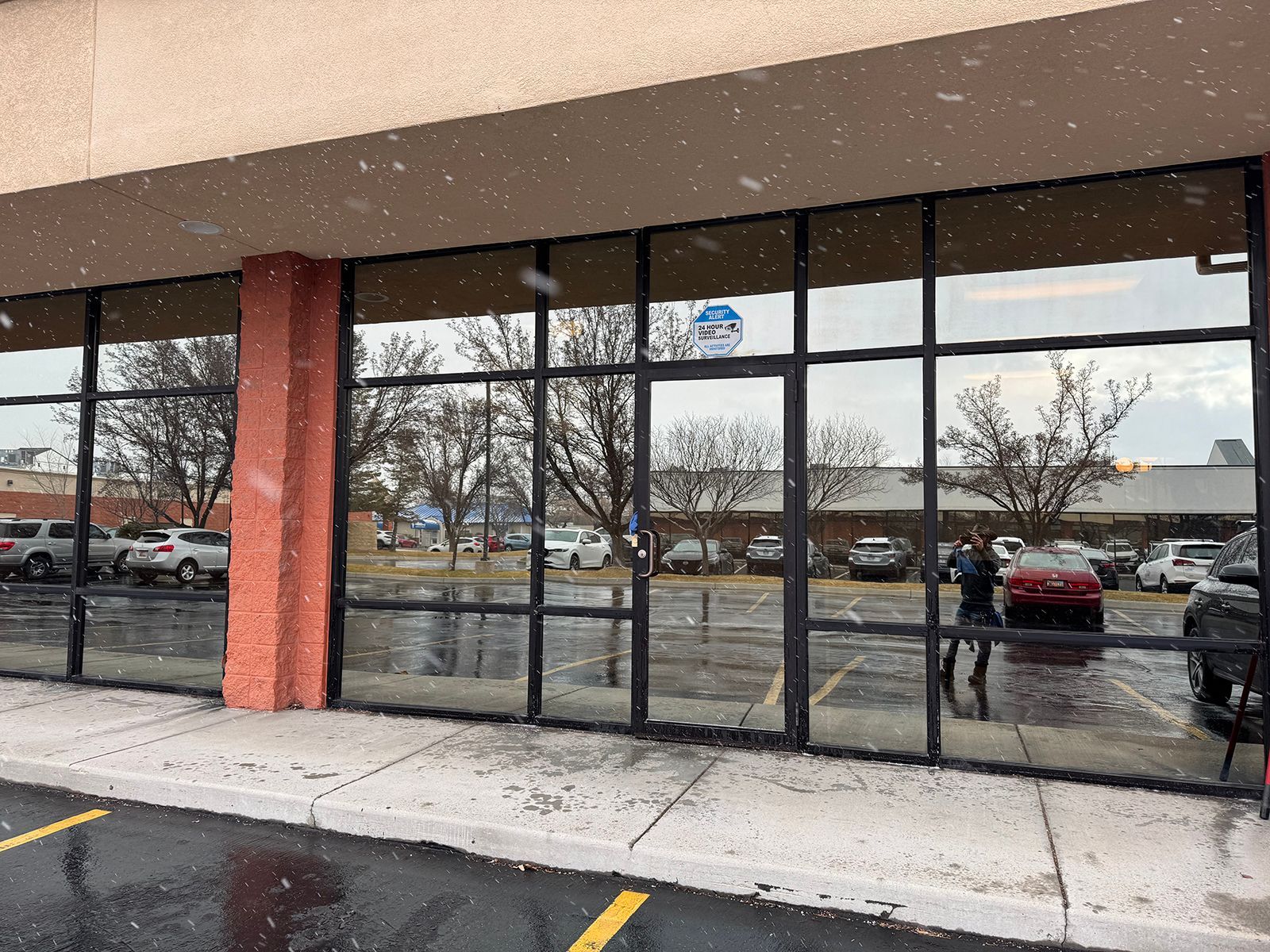 Building exterior with large windows reflecting a parking lot, trees, and falling snow.