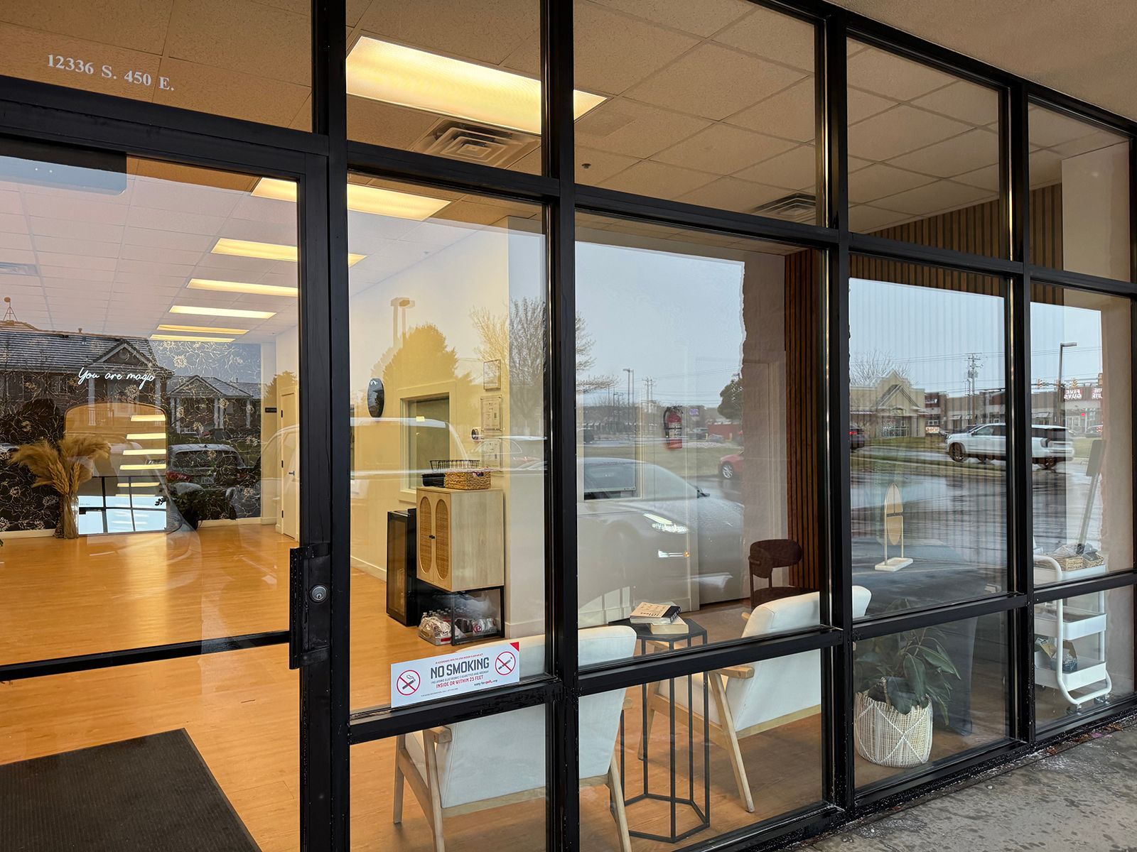 Exterior view of a commercial storefront with large windows; interior furnishings visible. Black frames, wet pavement.