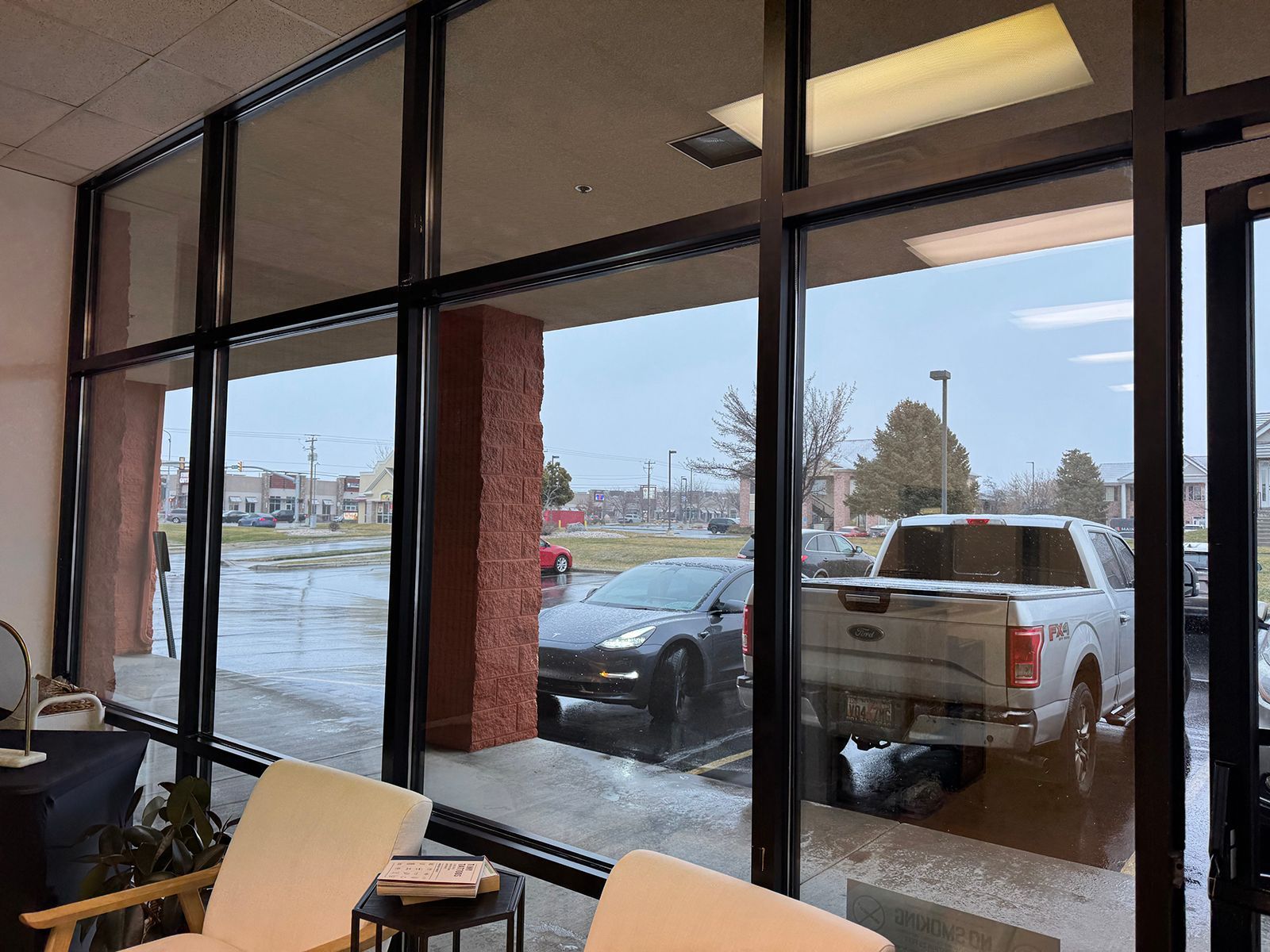 Interior view, large window overlooking a wet parking lot. Cars and a brick pillar are visible.