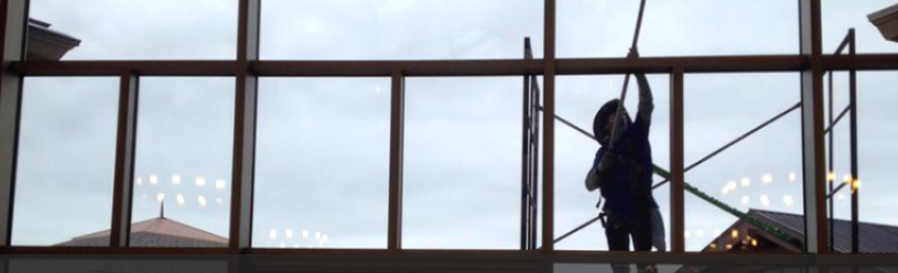 Person cleaning windows with a pole inside a building with large windows, overcast sky visible.
