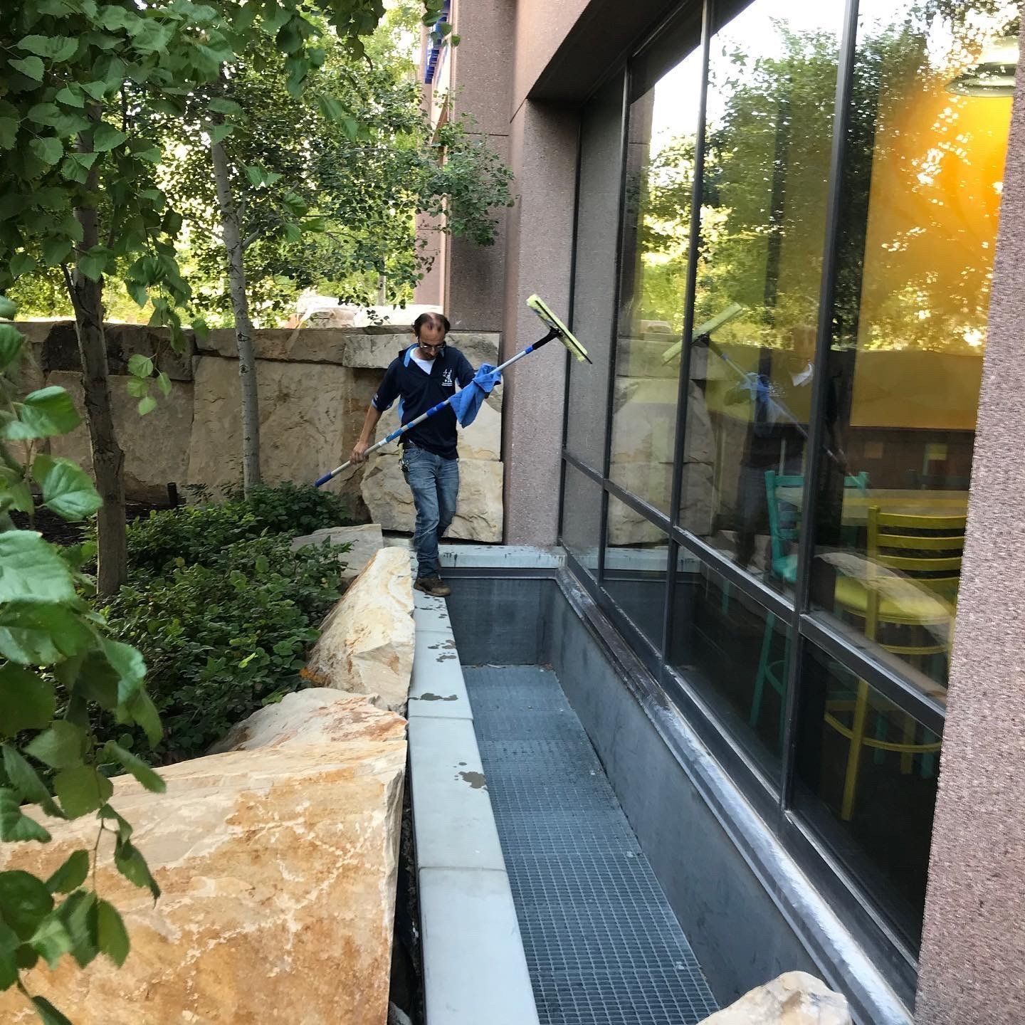 Man washing windows on a building with a squeegee; outdoors, near rocks and trees.