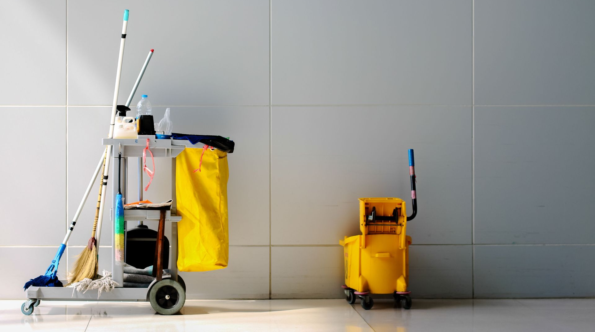 Cleaning supplies on carts: mops, brooms, buckets, and cleaning solutions against a tiled wall.