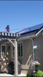 Person cleaning solar panels on a house roof with a long-handled brush on a sunny day.