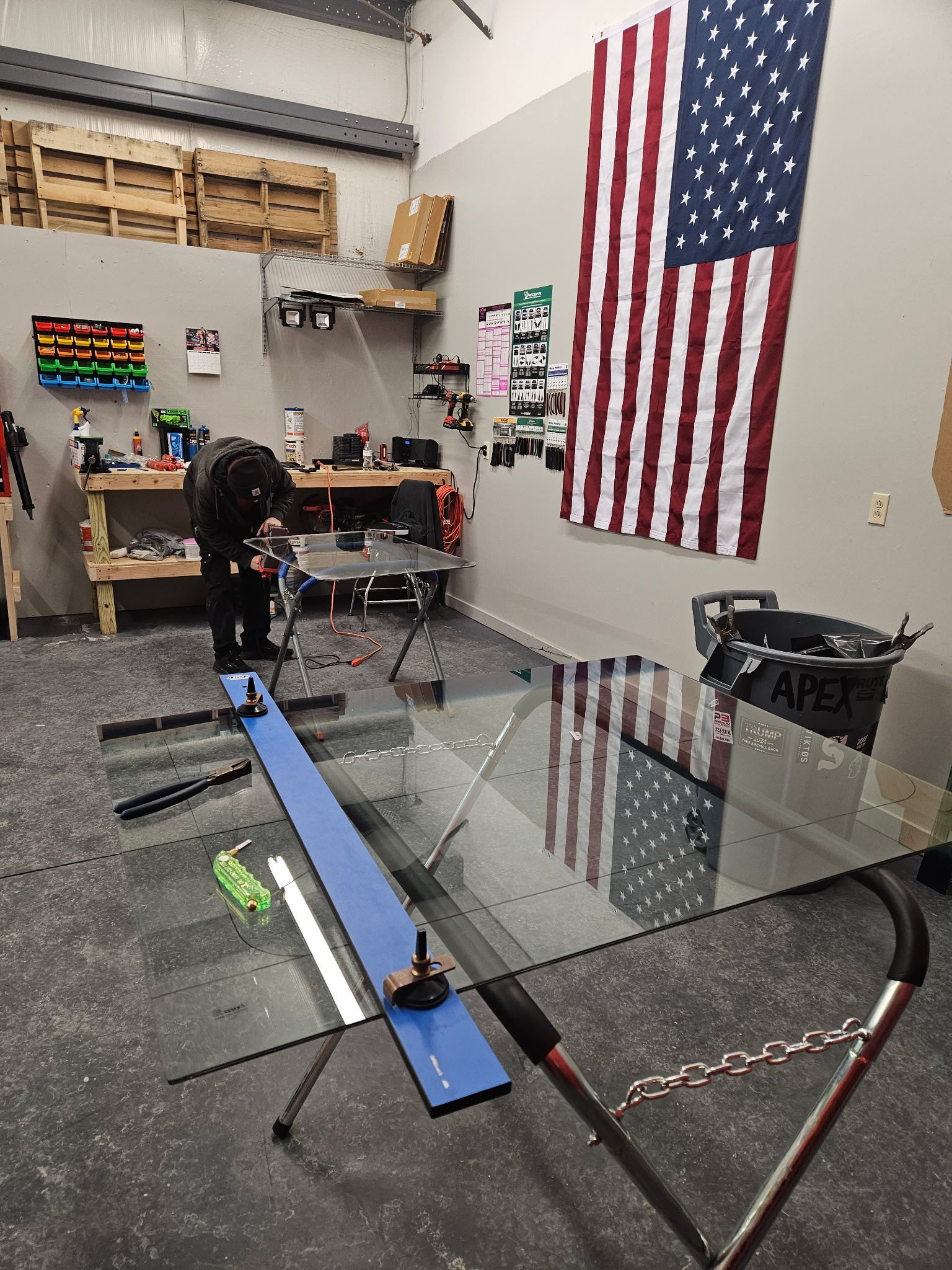 A man is working on a windshield in a garage with an American flag on the wall.