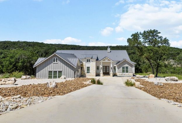 Stone house with a gray roof at the end of a driveway, surrounded by rocks and trees under a blue sky