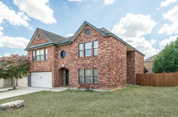 Two-story brick suburban house with a white garage, lawn, and wooden fence under a partly cloudy sky