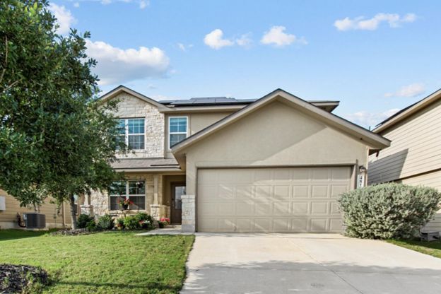 Two-story suburban house with beige siding, a two-car garage, and a front lawn under a blue sky.