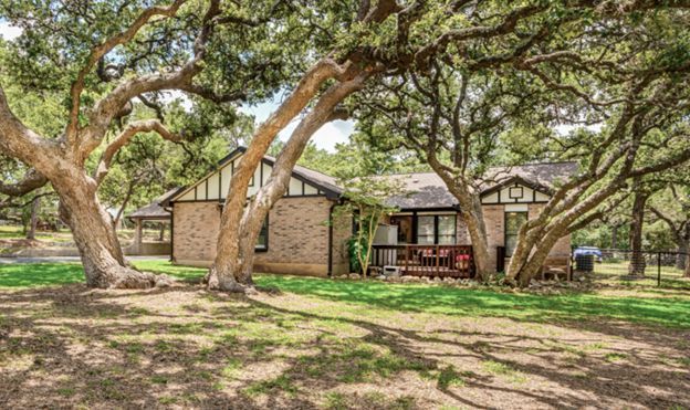 Rustic house under sprawling trees in a sunny yard with a porch and shaded lawn