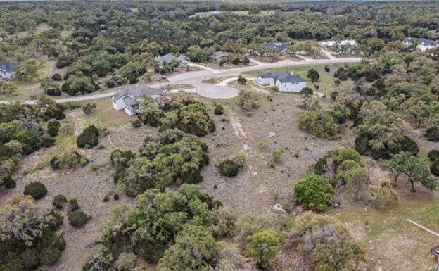 Aerial view of a rural property with a house, driveway, and scattered trees amid open scrubland.