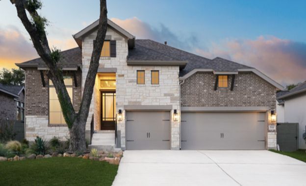 Modern two-story stone house with three-car garage at dusk, front porch lights on and tree in the yard