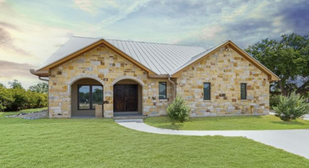 Stone house with arched front porch, light metal roof, and green lawn under a bright sky