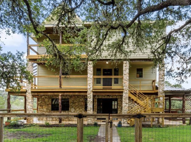 Two-story wooden house with wraparound porch and stairs, surrounded by large trees and a fenced yard.