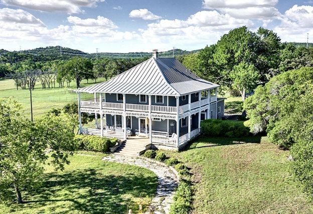 White gazebo-like house with wraparound porch in a lush green field under a cloudy sky