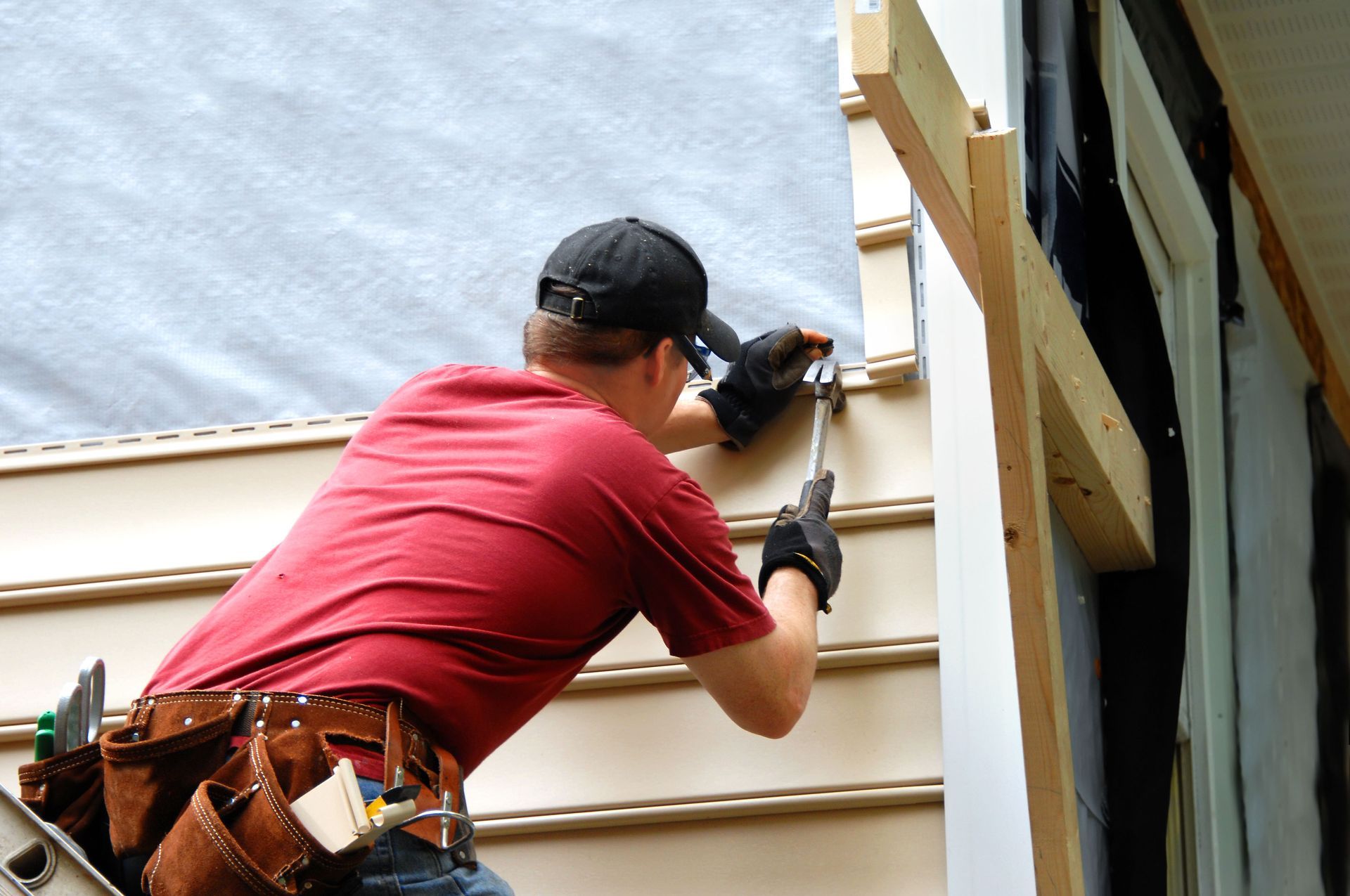 Construction worker installing siding on a building's exterior; wearing a red shirt, black hat, and tool belt.