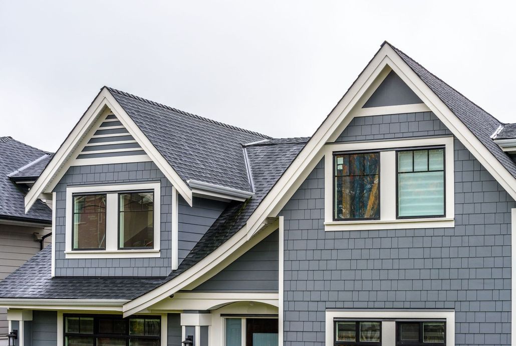 Gray-shingled house with white trim, two dormers with windows, against a cloudy sky.