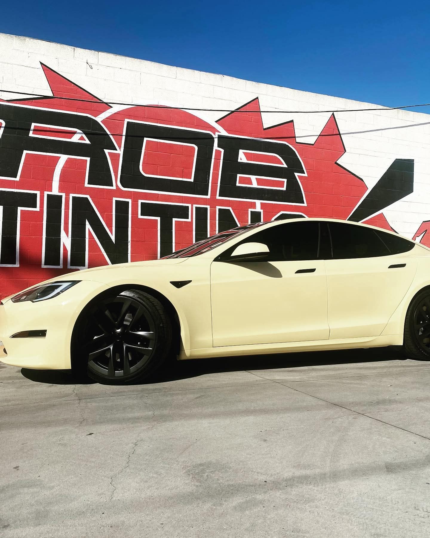 Cream-colored sports car parked in front of a red-and-black graffiti wall under a bright blue sky