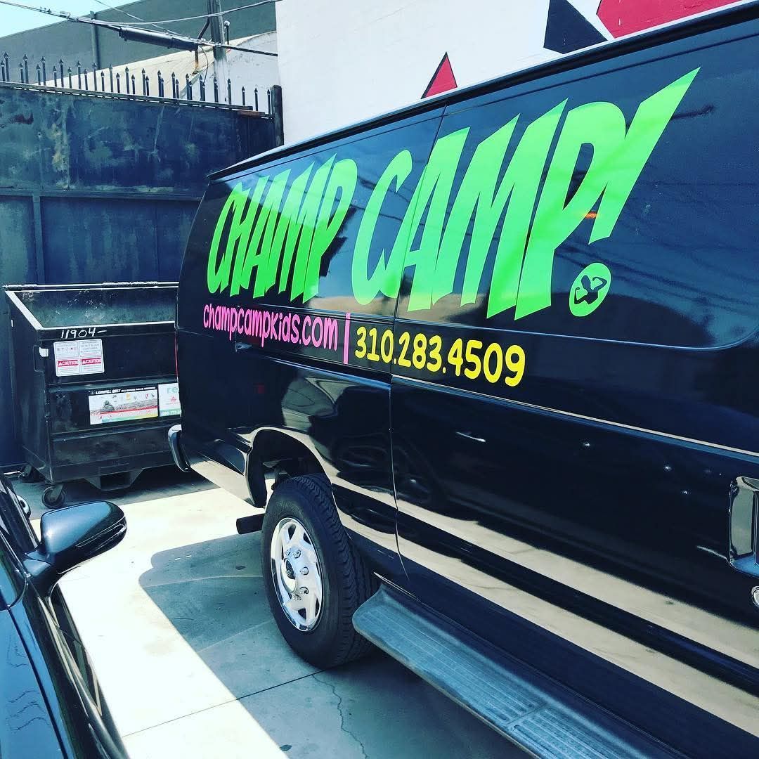 Black van with neon green “Camp” advertising parked outdoors in bright sunlight.