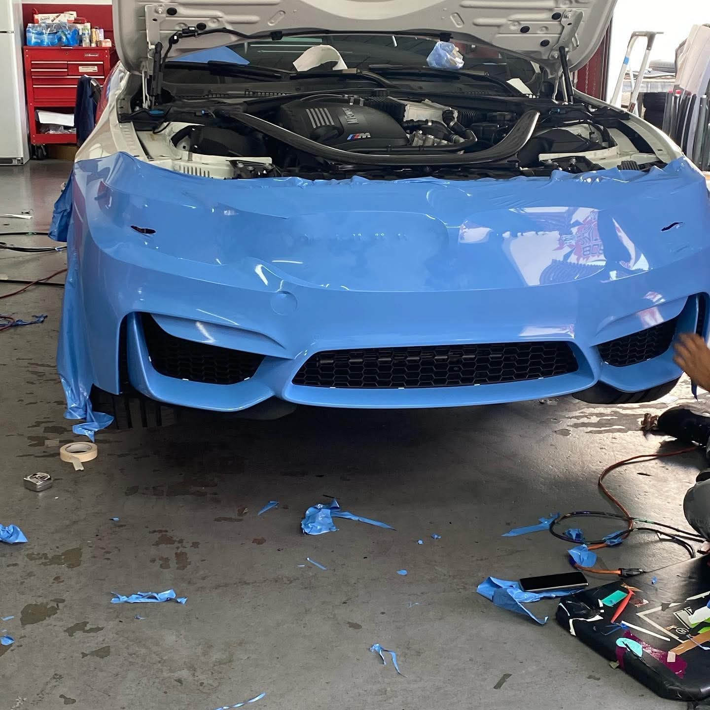 Damaged blue car front in auto repair shop with hood open and bumper wrapped in tape