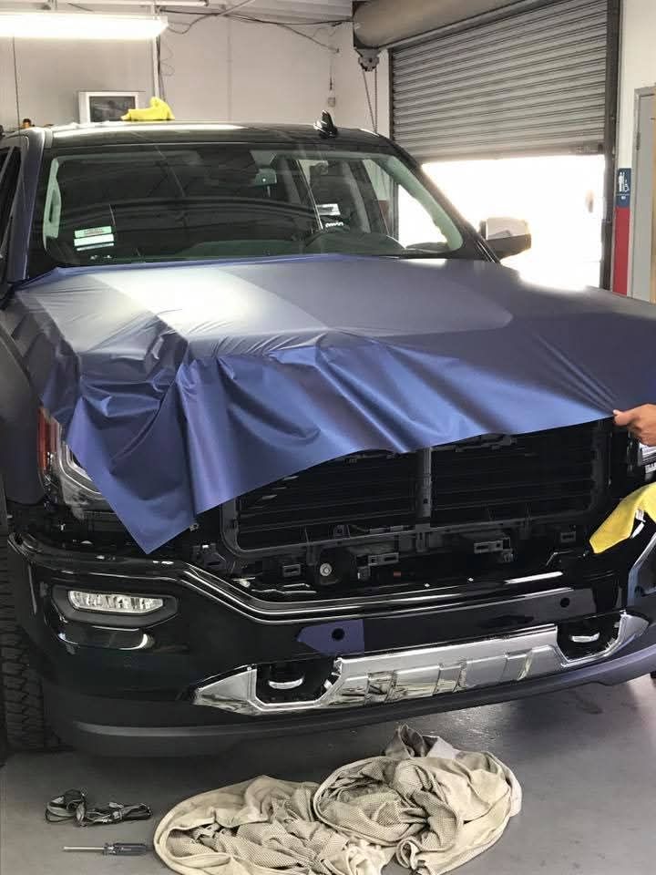 Black truck with front end damage in a garage, covered in blue protective film and repair materials nearby