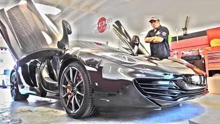 Black sports car in a garage with a person standing beside it, hood open and front doors raised.