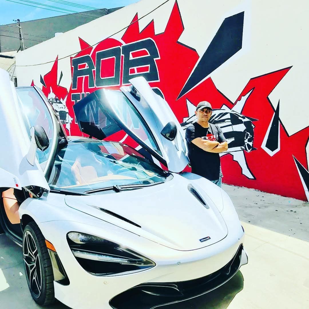 White sports car with gullwing doors open beside a graffiti wall, with a person holding a sign nearby