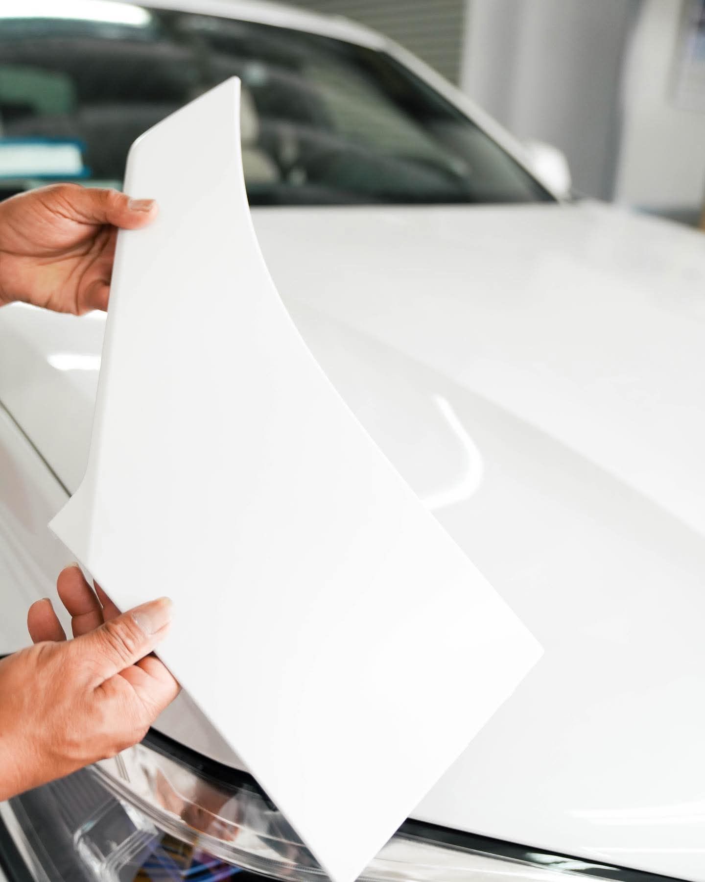 Hands peeling a white protective film from a white car hood in a garage.
