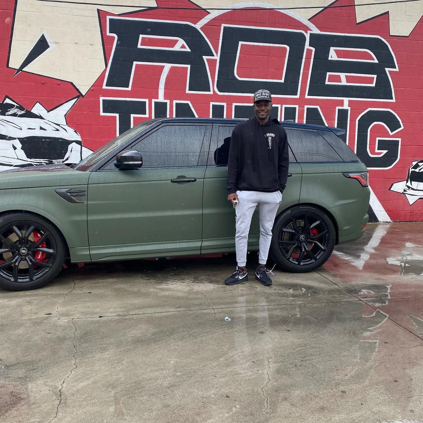 Man standing beside an olive green SUV in front of a red-and-black graffiti wall.