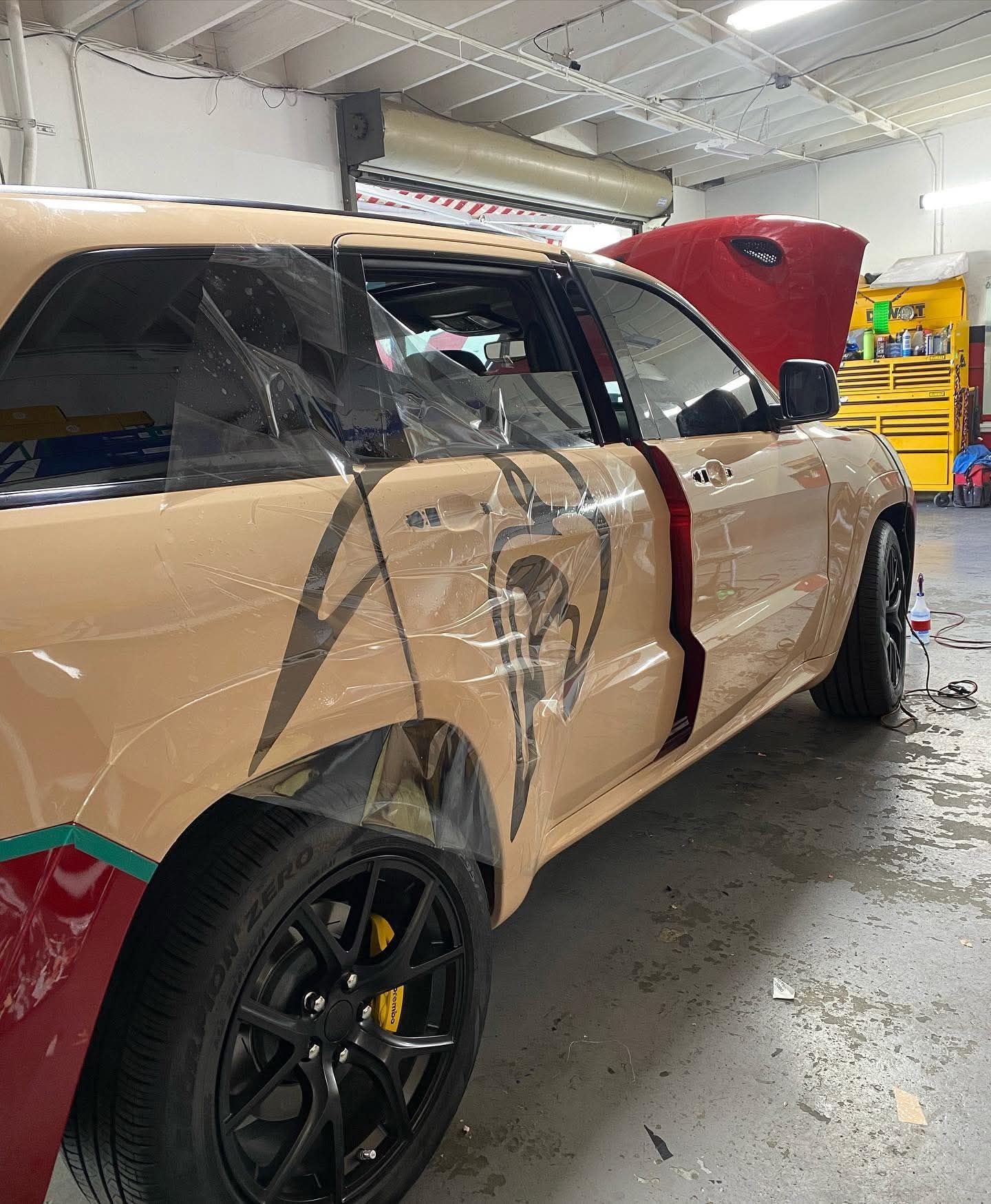 Damaged tan SUV in an auto shop, rear door crushed and hood open.