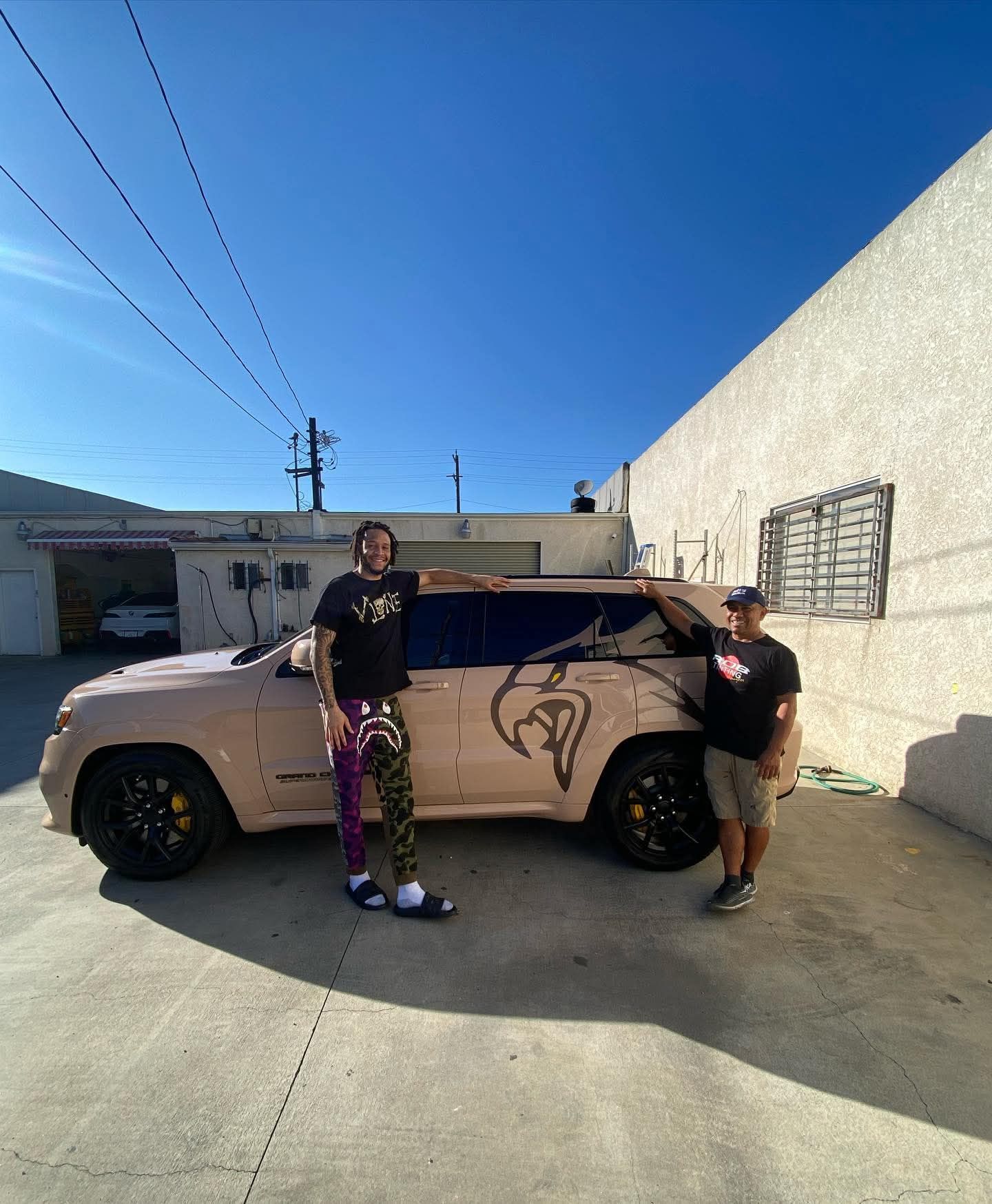 Two people posing beside a tan car in a sunny parking lot with a clear blue sky.