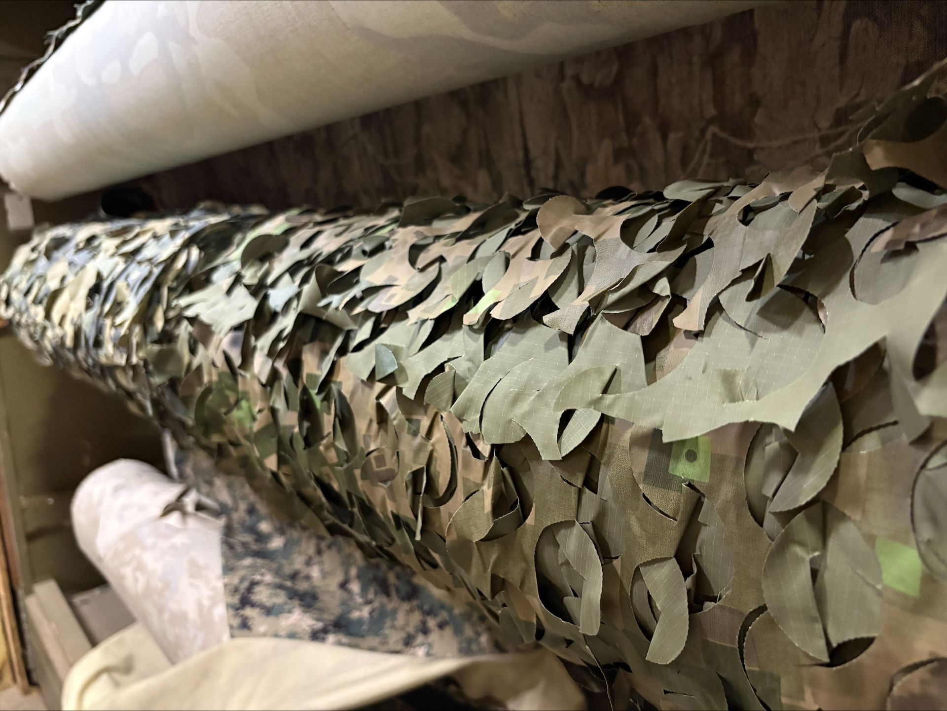 Roll of camouflage fabric with leaf-shaped cutouts, stacked with other fabrics in a storage area.