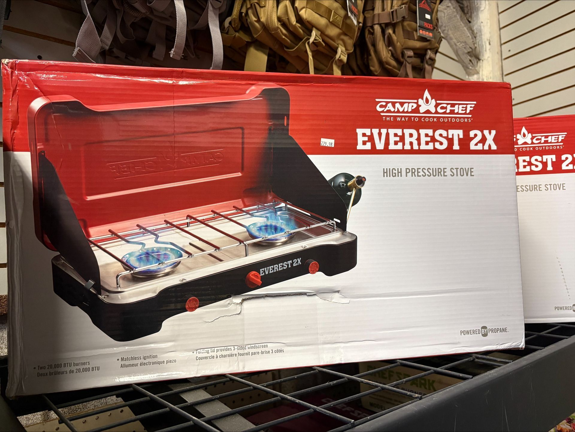 Boxed Camp Chef Everest 2X high-pressure stove on a store shelf, white with red accents, two burners visible.