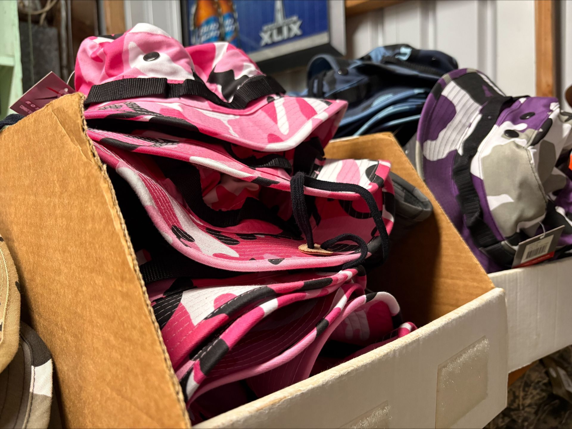 Box filled with folded pink camouflage bucket hats, other hats in background.