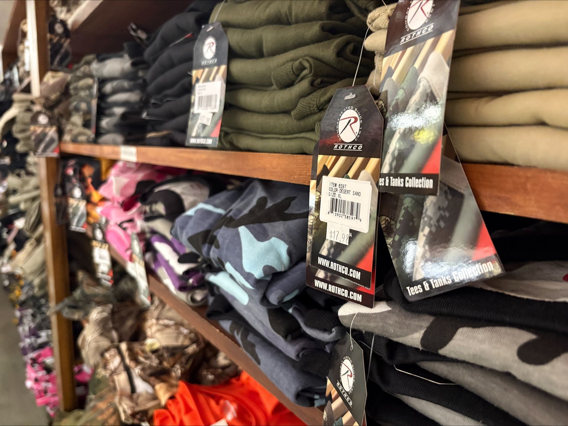 Shelves stocked with folded camouflage clothing in various colors, with price tags.
