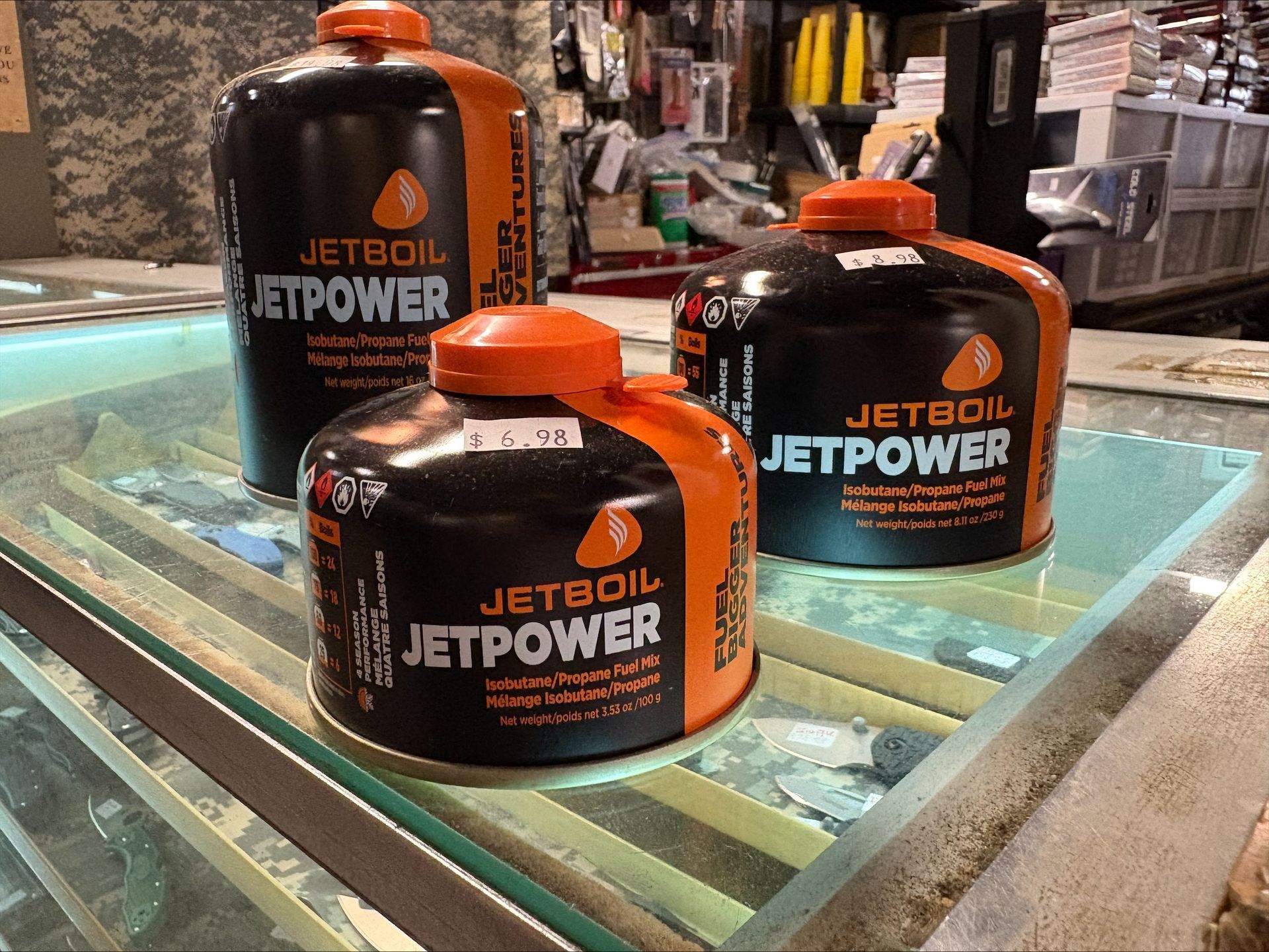 Three Jetboil Jetpower fuel canisters, black and orange, on a glass display case in a store.